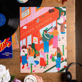 The Trevell Urban Stroll 500-piece eco-friendly puzzle features people outside a red record store. Vinyl records, headphones, and a coffee cup sit on a wooden table next to the completed puzzle.