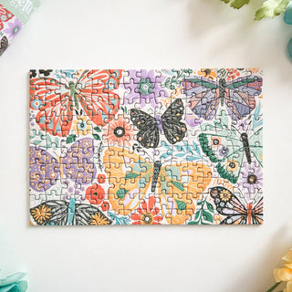 The Butterfly Bouquet | 150 Piece Jigsaw Puzzle by Spaghetti Tree features vibrant butterflies and flowers on a white background with leafy green and blue accents—a beautiful, nature-inspired puzzle.
