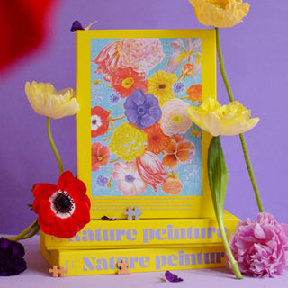 A vibrant floral print in a yellow frame stands on two yellow books against a purple background, surrounded by fresh flowers and pieces of the Jour Férié Nature Peinture 1,000 Piece Jigsaw Puzzle, made in France.