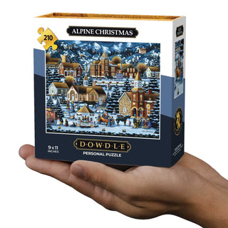 A hand holds a Dowdle Alpine Christmas | 210 Piece Personal Jigsaw Puzzle, featuring a festive winter village scene inspired by small town celebrations on the cover.