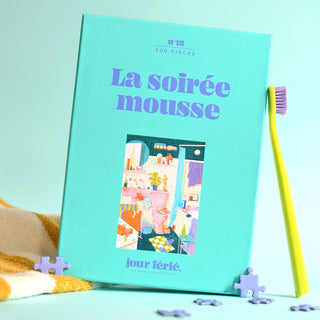 A turquoise box of the Jour Férié La soirée mousse 500 Piece Jigsaw Puzzle stands upright beside a purple toothbrush and yellow-striped towel, with two loose ribbon-cut pieces nearby. Made in France.