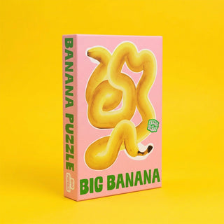 The Big Banana | 500 Piece Jigsaw Puzzle by Cloudberries features a unique banana-shaped design with a large, twisted banana on a pink and yellow background—perfect for fans of eye-catching puzzles.