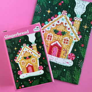 A Gingerbread | 99 Piece Jigsaw Puzzle box next to its partially assembled ribbon-cut puzzle, both depicting a decorated gingerbread house on a dark green background with holly and stars.