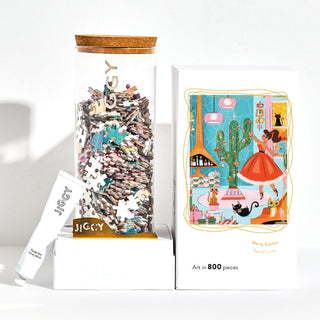 A tube, a jar with puzzle pieces, and a box featuring the Merry Cactus artwork—a woman in a red dress, cats, and plants—highlight this Art in 800 Pieces JIGGY Puzzles jigsaw.