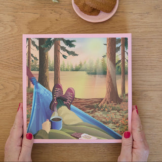 A person with red nail polish holds the ViSSEVASSE "Hiking & Me" 500 piece FSC certified puzzle, featuring art of someone relaxing in a hammock by a lake with coffee, chocolate, sunset views, and cookies on a table above.