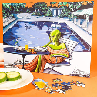 The Sunset Sector Seven 1,000 piece jigsaw puzzle by Chuffed Puzzles features surreal art by Heike Blakley of a green alien relaxing poolside with snacks; completed puzzle shown with pieces and cucumber slices in front.