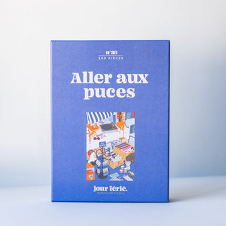 The Jour Férié Aller Aux Puces | 500 Piece Jigsaw Puzzle comes in a blue box and features a vibrant flea market scene. This charming puzzle is proudly made in France.