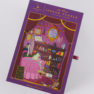 A jigsaw puzzle box from Good Juju Ink displays an illustrated fortune teller’s room filled with a cat, mystical objects, and shelves of books. The purple magnetic keepsake box is labeled “500 Piece Jigsaw Puzzle” and “Madame Oddstrich,” accented with dazzling gold foil details for an enchanting touch.