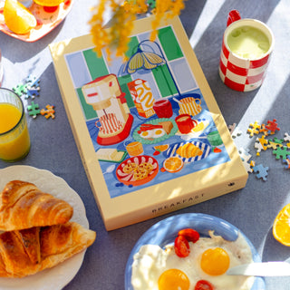 A breakfast scene featuring croissants, eggs, juice, a green drink, and yogurt with tomatoes beside Das Puzzle Kollektiv's "Frühstück | 1,000 Piece Jigsaw Puzzle" box on a table scattered with puzzle pieces.