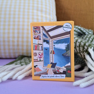 The Hytte Les Pieds Dans L'eau 100 Piece Jigsaw Puzzle by Pièce Rapportée features a cozy room with a seaside view on its yellow box—ideal for travel journals or mini puzzles—shown on a green checked blanket and beige plaid cushion.