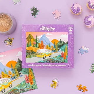 The Road Trip | 250 Piece Jigsaw Puzzle by Villager Puzzles features a vibrant van and mountain scene—perfect as travel-themed decor or a fun road trip activity. Puzzle pieces, cupcakes, and a drink appear on the colorful purple box surface.