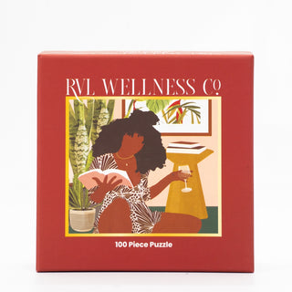 The Wine Down 100 Piece Jigsaw Puzzle by RVL Wellness Co. features a red box with art of a woman reading and sipping a drink among plants and framed pictures—perfect for relaxation and mindfulness.