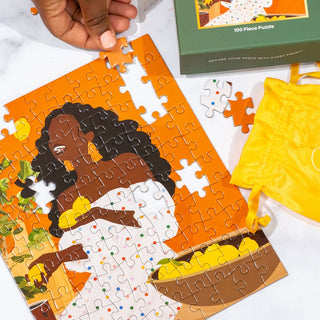 A hand places a piece into RVL Wellness Co.’s “This Kitchen Is For Laughing” 100-piece puzzle, showing a woman with curly hair and a bowl. Yellow pouch and box nearby—perfect for self-care or relaxing mini puzzle time.