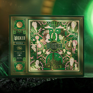 The theory11 Wicked | 1,000 Piece Jigsaw Puzzle comes in a decorative box with Emerald City artwork, illustrated characters, a hot air balloon, and green-gold accents. Suitable for ages 14+.