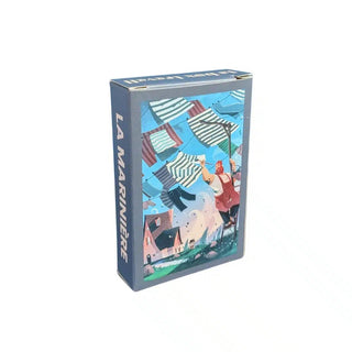 A box labeled La Marinière | 99 Piece Jigsaw Puzzle by Trevell features an illustrated scene of a person hanging Breton striped shirts on a clothesline above houses in a breezy coastal village, capturing the charm of Trevell in this delightful mini puzzle.