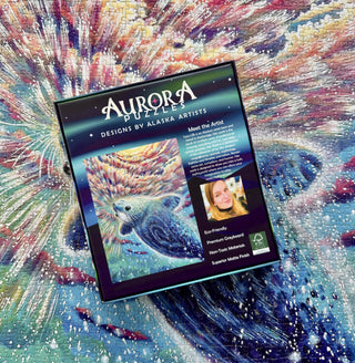 A box of Aurora Puzzles’ The Selkie | 1,000 Piece Jigsaw Puzzle rests on a completed, vibrant puzzle featuring Tamara Ridgway’s otter artwork, resembling an abstract aurora or explosion.