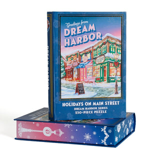 A Kingdom of Threads Dream Harbor Holidays On Main Street 250-piece jigsaw puzzle is presented in an upright, book-shaped keepsake box with the bottom half lying flat, featuring a festive street scene from the Dream Harbor Series—an ideal display piece for your cozy winter puzzle collection.