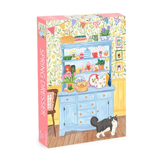 The Spring Dresser 500 Piece Jigsaw Puzzle by Olleo Puzzles features a charming blue dresser with dishes, flowers, and decorations set against wallpaper, plus a black and white cat on the wooden floor.