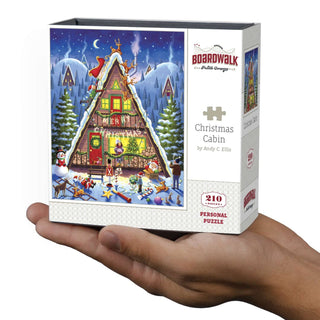 A hand holds a box of Boardwalk Puzzle Company's Christmas Cabin | 210 Piece Personal Jigsaw Puzzle, a festive holiday scene perfect for fans of cartoon characters and Christmas fun.
