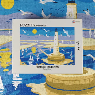 The completed Cordouan Lighthouse | 1,000 Piece Jigsaw Puzzle by Aparté displays a lighthouse on an island with sailboats, seagulls, and a bright sky; the box with the same image rests on top.