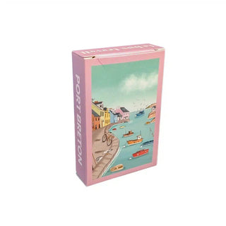 A rectangular box with pink edges featuring an illustration of a seaside village, boats in the water, and houses along the shore. The side reads PORT BRETON. The Trevell Port Breton | 99 Piece Jigsaw Puzzle is a charming mini harbor puzzle, perfect for anyone who enjoys coastal scenes.