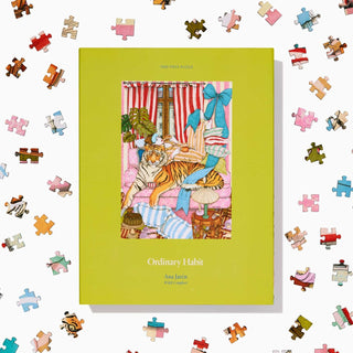 A yellow box of the Wild Comfort | 1,000 Piece Jigsaw Puzzle by Ordinary Habit, featuring cat-themed artwork, sits among colorful scattered pieces on a white background.