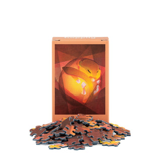 A box of the Harvest Mouse | 99 Piece Mini Jigsaw Puzzle by Happily Jigsaw Puzzles, featuring an abstract orange and yellow illustration and made from recycled board, stands behind a scattered pile of puzzle pieces on a white background.
