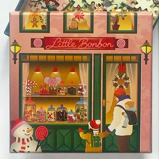 Delight in the charming scene of a candy shop called Little Bon Bon, featuring jars of sweets in the window, a snowman holding a lollipop, and an adult with two children at the door. The ViSSEVASSE "Little Bon Bon | 150 Piece Jigsaw Puzzle" perfectly captures this whimsical moment, making it an ideal puzzle for candy lovers of all ages.