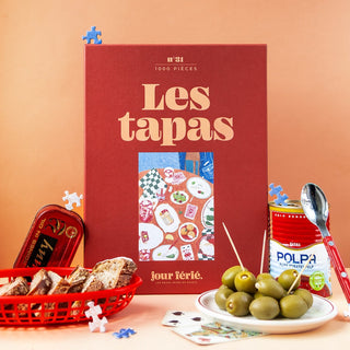 A red Jour Férié Les tapas | 1,000 Piece Jigsaw Puzzle box is set among canned sardines, bread, green olives, a spoon, a can of tomato polpa, and loose ribbon cut pieces. Made in France.