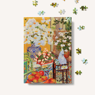 The Puzzle Theory’s “Mangos and Matilijas” is a 500-piece eco-friendly puzzle showcasing a colorful still life with white Matilija poppies, fruit, a blue vase, and a bird statue on a patterned table, with loose pieces pictured around the box.