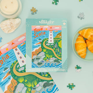The Nova Scotia Shores | 500 Piece Jigsaw Puzzle by Villager Puzzles rests on a light blue surface, with loose pieces, a candle, seashell, another puzzle, and croissants—a charming Nova Scotia souvenir and coastal decor accent.