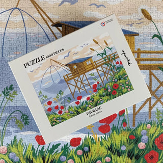 A Pauillac | 1,000 Piece Jigsaw Puzzle by Aparté, featuring Paola Hirou’s vibrant seaside house on stilts, sits atop the completed puzzle and highlights the same stunning, floral coastal scene.