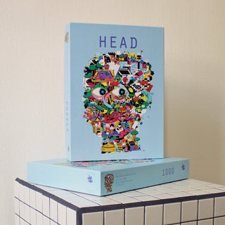 The Das Puzzle Kollektiv Head | 1,000 Piece Jigsaw Puzzle is a plastic-free box featuring a vibrant illustration of objects and characters forming a head, shown atop its matching box on a tiled surface.