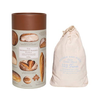 A cylindrical box labeled "Sourdough | 500 Piece Jigsaw Puzzle" with bread images holds an artisan puzzle next to a 123 Farm drawstring bag printed with farm-themed text.