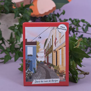 Pièce Rapportée’s Dans Les Rues De Bergen 100 Piece Jigsaw Puzzle comes in a colorful box featuring Bergen’s street scene with buildings, flags, flowers, and the title—perfect as a travel-themed mini puzzle or unique gift.