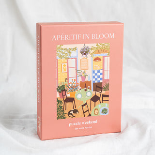 Puzzle Weekend’s Apéritif in Bloom | 500 Piece Jigsaw Puzzle features a charming illustrated European café scene with tables, chairs, flowers, and a cat—perfect for any puzzle lover.
