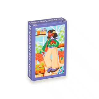 The Trevell Le Magasin Des Fleurs 99 Piece Jigsaw Puzzle features a colorful box with art of a person holding flowers by a window. A perfect mini challenge for flower shop puzzle fans!.