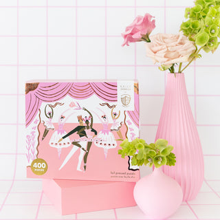 The Pirouette "Stage" 400-piece jigsaw puzzle by Jollity & Co. features a ballerina design with rose gold foil accents, displayed on a pink pedestal next to a pink vase and flowers, set against a pink grid background.