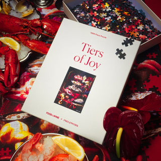 A Piecework Puzzles Tiers of Joy | 1,000 Piece Jigsaw Puzzle box sits on a table with seafood dishes, lemon slices, and a partially completed puzzle—all made from recycled paper.