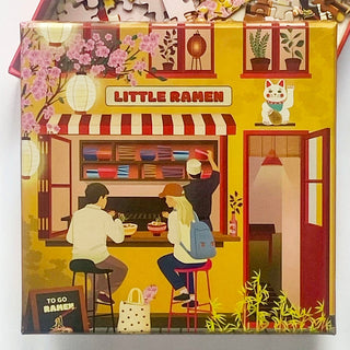 Two friends enjoy steaming ramen at the outdoor counter of Little Ramen as a chef works inside; cherry blossoms, lanterns, and a beckoning cat add delightful touches. This inviting scene is beautifully captured in the Little Ramen | 150 Piece Jigsaw Puzzle by ViSSEVASSE.