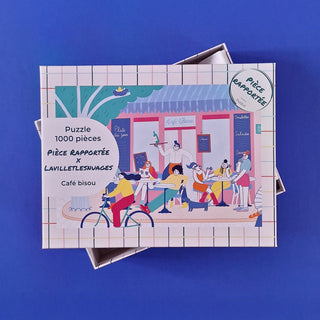 The Café Bisou | 1,000 Piece Jigsaw Puzzle by Pièce Rapportée features an illustrated café scene with outdoor diners, a passing cyclist, and pastel hues set against a blue background.