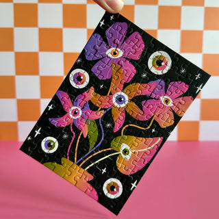 A hand displays the Surreal | 99 Piece Jigsaw Puzzle by Puzzledly, featuring colorful, eye-centered flowers on black. The pocket-sized puzzle has playful ribbon-cut pieces; orange and white checks decorate the wall behind.
