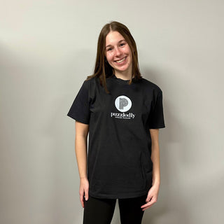 A woman stands against a plain light gray wall, smiling in a black Puzzledly T-Shirt by Puzzledly, paired with black pants.