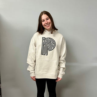A person with straight brown hair, smiling at the camera, models the Puzzledly "Piece Your Sh*t Together" Hoodie—a cream cotton fleece pullover featuring a bold black geometric P—against a plain light gray wall.