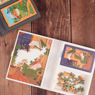 Two Trevell Puzzle Book cases with partially completed jigsaw puzzles are shown on a wooden surface. Pieces are sorted in compartments, and a mini puzzle accessory is placed beside The Puzzle Book.