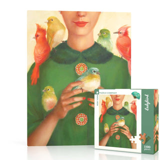 A woman in a green dress holds a blue bird while colorful birds perch on her arms. The scene from Janet Hill’s art appears on the Ladybird Mini | 100 Piece Jigsaw Puzzle by New York Puzzle Company in the foreground.