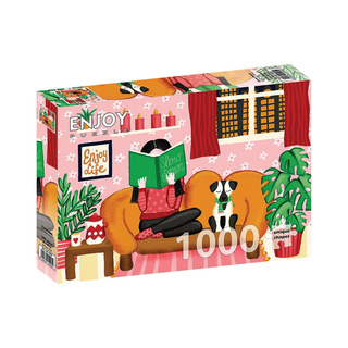 The ENJOY Slow Down 1,000 Piece Jigsaw Puzzle features a cozy, pink-themed scene of someone reading on a couch with a dog, surrounded by plants and decor. Enjoy the satisfying challenge of this ribbon cut puzzle.