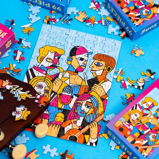 The Minizz Good Song 99 Piece Jigsaw Puzzle, featuring colorful geometric cubist figures, is completed on a blue surface with puzzle pieces and boxes nearby.