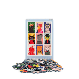 A box of the Art Cats | 99 Piece Mini Jigsaw Puzzle by Happily Jigsaw Puzzles—nine stylized felines depicted in vibrant artwork, with puzzle pieces playfully scattered in front.