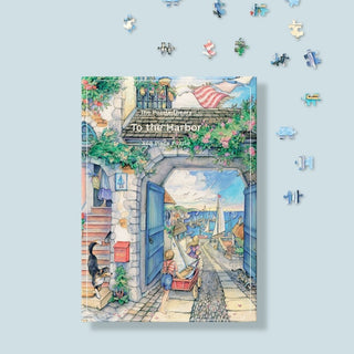 The Puzzle Theory’s “To the Harbor” is an 896-piece jigsaw puzzle illustrated by Kim Jacobs, featuring a charming seaside village scene with people, boats, and flowers; eco-friendly pieces are scattered on a light blue surface.